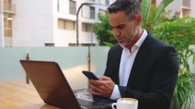 Takım elbiseli yetişkin bir adam elinde bir telefon tutuyor, mesajlar yazıyor ve internetten bir uygulama kullanarak haberleri okuyor.