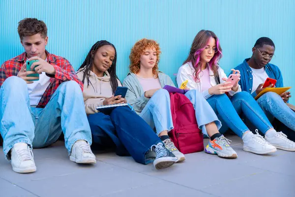 Beş genç öğrenciden oluşan çeşitli bir grup yerde oturup, mavi bir arkaplana karşı akıllı telefonlarıyla gülümsüyor ve etkileşime geçiyorlar..