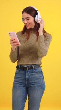 Dikey video genç beyaz kadın sarı arka planda cep telefonuyla müzik dinleyip dans ediyor. İnsan duyguları kavramı, yüz ifadesi. İlanınız için boşluk kopyala