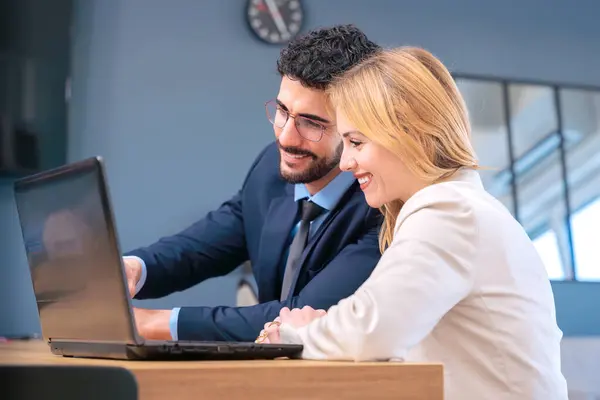 Modern bir ofiste iş birliği yapan iki iş arkadaşı dizüstü bilgisayarda çalışırken gülümsüyor. Profesyonel ama dost canlısı bir ortamda fikirlerini paylaşıyorlar, üretkenliği ve takım çalışmasını teşvik ediyorlar..