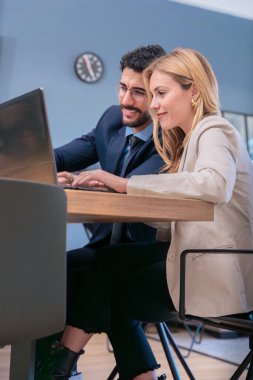 Modern bir ofiste iş birliği yapan iki iş arkadaşı dizüstü bilgisayarda çalışırken gülümsüyor. Profesyonel ama dost canlısı bir ortamda fikirlerini paylaşıyorlar, üretkenliği ve takım çalışmasını teşvik ediyorlar..