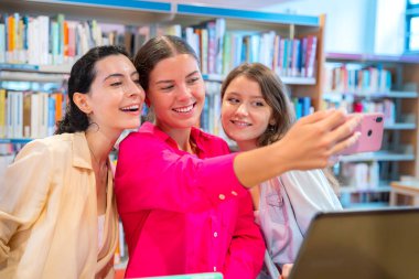 Kütüphane ortamında selfie çekerken gülümseyen üç kadın. Kitap raflarıyla çevrelenmiş bir şekilde, bir neşe, arkadaşlık ve ders arası yakalıyorlar, pozitif öğrenme ortamını yansıtıyorlar.