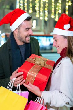  Noel Baba şapkası takan neşeli çift bayram gecesinde hediyelerini değiş tokuş ediyor. Bayram ışıklarıyla çevrili, sarılı hediyeler ve renkli alışveriş çantaları taşıyorlar. Şehirde Noel 'in keyfini kutluyorlar.