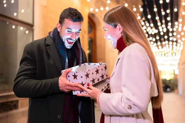 Heyecanlı çift, bayram ışıklarıyla çevrili şenlikli bir şehir gecesinde parıldayan bir hediye kutusu açıyor. Noel, hediye verme, kutlama ve neşeli bayram temaları için mükemmel.