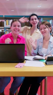 Üç genç kız öğrenci bir kütüphanede birlikte çalışıyorlar, bir dizüstü bilgisayar kullanıyorlar ve kamerayla uğraşıyorlar. Odaklanmış ama dost canlısı görünüyorlar, sessiz bir akademik ortamda bir projede işbirliği yapıyorlar.