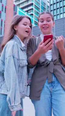 İki mutlu genç kadın dışarıdayken akıllı telefonlarından heyecan verici haberler alıyor. Biri şaşırmış bir yüz ifadesi sergilerken, diğeri içten bir şekilde gülümsüyor, doğadaki neşeli bir anı yakalıyor.