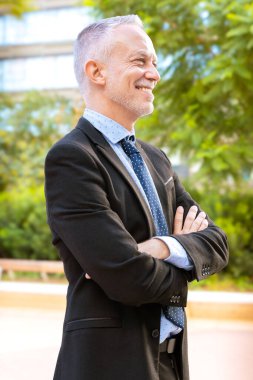 Takım elbiseli ve kravatlı gülümseyen kıdemli işadamı, kolları bağlı güvenle ayakta duruyor. Doğal bir açık hava ortamında yakalanmış, dışarı çıkma deneyimi, profesyonellik ve pozitiflik..