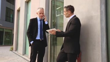 Takım elbiseli iki işadamı modern bir ofis binasının dışında iş konuşuyorlar. Biri telefon kullanırken, diğeri iş ortamında iletişim ve takım çalışmasını vurgulayan bir tablet taşıyor.