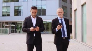  Takım elbiseli iki işadamı akıllı telefon kullanırken şehir merkezinde yürüyorlar. Profesyonel bir şirket ortamında iletişim teknolojisi ve üretkenliği temsil ediyorlar.