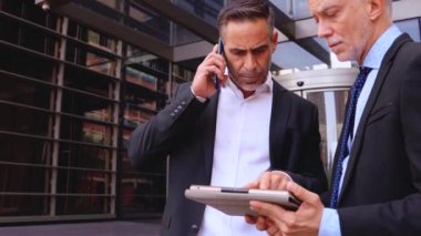 İki iş adamı modern bir ofis binasının dışında çalışmayı tartışıyor. Biri telefonla konuşurken, diğeri tabletteki bir belgeyi inceliyor. Şirket kentsel ortamında profesyonel takım çalışması