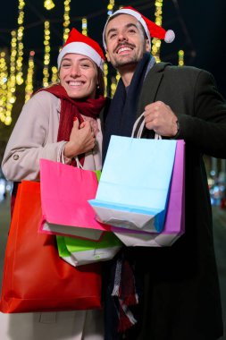 Noel Baba şapkası takan, Noel ışıklarının altında duran gülümseyen bir çift. Birbirlerine sıcak bir şekilde bakıyorlar, neşeli bir tatil ortamı yaratıyorlar. Birinin telefonu var. Dikey.