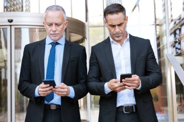 Ofis binasının önünde duran takım elbiseli iki profesyonel iş adamı ikisi de akıllı telefonlarına odaklanmış. İş ortamlarında teknoloji kullanımı, şirket hayatı ve modern iletişimi temsil eder.