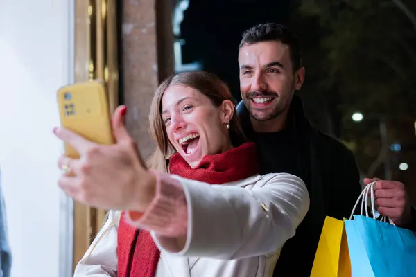 Şenlikli bir şehir gecesinde ellerinde renkli alışveriş torbalarıyla selfie çeken gülümseyen bir çift. Noel, teknoloji, alışveriş ve bayram kutlamaları için mükemmel
