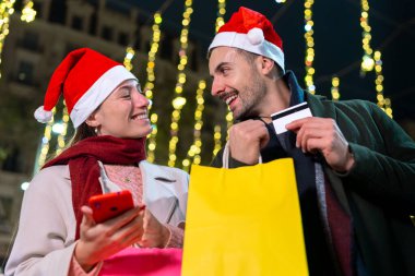  Noel Baba şapkası takan, Noel ışıklarının altında duran gülümseyen bir çift. Birbirlerine sıcak bir şekilde bakıyorlar, neşeli bir tatil ortamı yaratıyorlar. Biri telefonu tutuyor, anıları yakalıyor..