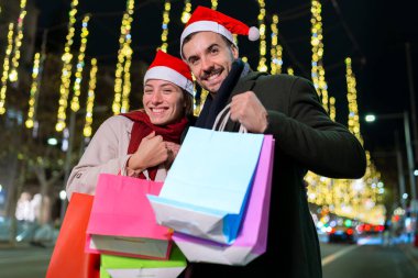 Noel Baba şapkası takan, Noel ışıklarının altında duran gülümseyen bir çift. Birbirlerine sıcak bir şekilde bakıyorlar, neşeli bir tatil ortamı yaratıyorlar. Birinin telefonu var..