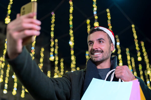 Мужчина в шляпе Санты улыбается, делая селфи и держа сумки на праздничной улице города, освещенной светящимися праздничными светильниками. Идеально подходит для Рождества, технологий и праздников