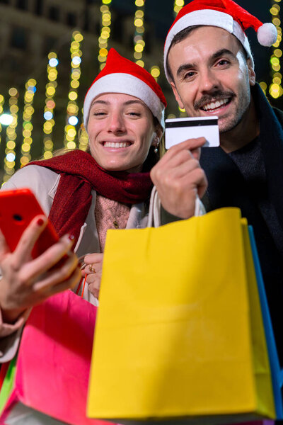 Улыбающаяся пара в шляпах Санты, стоящая под праздничными рождественскими огнями. Они смотрят друг на друга тепло, создавая радостную праздничную атмосферу. Один держит телефон. Вертикальный.