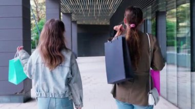 Başarılı bir alışveriş çılgınlığından sonra ellerinde renkli alışveriş torbalarıyla sokakta yan yana yürüyen kadınlar. Sıradan giyiniyorlar, kot pantolon ve spor ayakkabı giyiyorlar, açık hava alışverişinin tadını çıkarıyorlar.