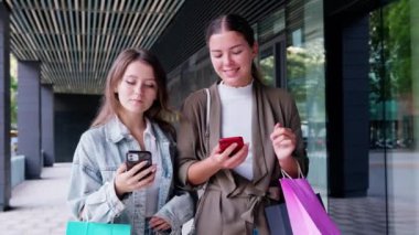 İki genç kadın renkli alışveriş torbaları taşıyarak bir alışveriş bölgesinde yürüyorlar. Bir kadın telefonunu tutarken diğeri telefonunu kontrol ediyor, gülümsüyor ve alışveriş gününün tadını çıkarıyor..