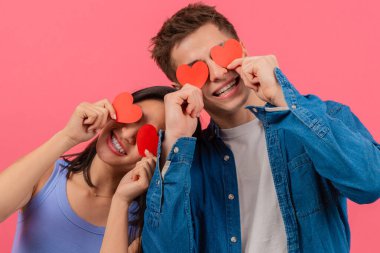 Gülümseyen bir çift canlı pembe bir arka planda gözlerinin üzerinde kırmızı kalp şekilleri tutuyor. Görüntü, sevgililer günü ya da romantik temalar için mükemmel bir sevgi, neşe ve eğlence sunar..