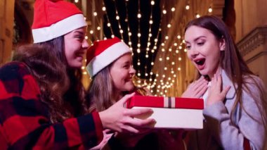 Noel Baba şapkası takan üç kadın, festival ışıklarıyla süslenmiş bir caddede gülüp hediyelerini değiş tokuş ediyorlar. Neşeli an, tatil sezonunun ve arkadaşlığın ruhunu yakalar..