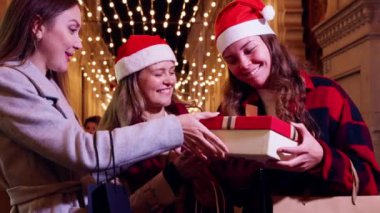 Noel Baba şapkası takan üç kadın, festival ışıklarıyla süslenmiş bir caddede gülüp hediyelerini değiş tokuş ediyorlar. Neşeli an, tatil sezonunun ve arkadaşlığın ruhunu yakalar..