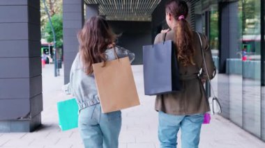 Başarılı bir alışveriş çılgınlığından sonra ellerinde renkli alışveriş torbalarıyla sokakta yan yana yürüyen kadınlar. Sıradan giyiniyorlar, kot pantolon ve spor ayakkabı giyiyorlar, açık hava alışverişinin tadını çıkarıyorlar.