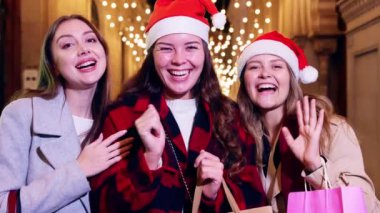 Noel Baba şapkası takan üç neşeli kadın gülüyor ve Noel gecesini kutluyor, ellerinde hediyeler ve şehrin şenlikli ışıklarının önünde poz veriyorlar. Neşeli, coşkulu bir arkadaşlık ve tatil ruhu anı..