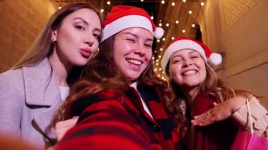 Noel Baba şapkası takan üç neşeli kadın gülüyor ve Noel gecesini kutluyor, ellerinde hediyeler ve şehrin şenlikli ışıklarının önünde poz veriyorlar. Neşeli, coşkulu bir arkadaşlık ve tatil ruhu anı..