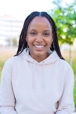  A close-up portrait of a joyful young woman with braided hair. She smiles brightly outdoors in a natural setting, with a soft background of greenery. The image captures positivity.Vertical clipart