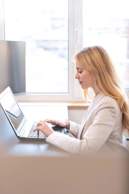 A businesswoman sits at a desk in a bright office, typing on a laptop. She is focused on her work, with natural light coming through the window, creating a professional and productive atmosphere.  clipart