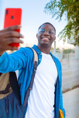  Genç bir adam açık havada sarı bir klasörü tutarken selfie çekerken parlak bir şekilde gülümsüyor. Gözlük takıyor ve sıradan bir kot ceket giyiyor. Arka planda çitleri ve yeşilliği olan güneşli bir gün var..