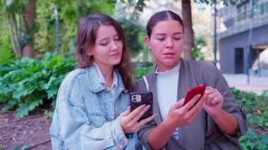 Heyecanlı iki genç kadın canlı bir şehir ortamında, akıllı telefonlarına bakarken heyecanlarını paylaşıyorlar. Sahnede renkli modern binalar, yayılan enerji ve bağlantı. 