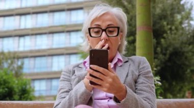 Gözlüklü ve spor ceketli yaşlı bir kadın parkta akıllı telefonuna bakarken şaşırıyor. Yeşillik ve modern bir bina dinamik bir ortam yaratır..