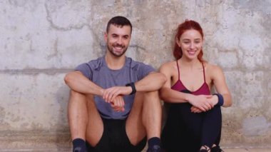 Formda bir çift yerde oturuyor, antrenmandan sonra birbirlerine gülümsüyorlar. Spor giysileri içinde beton bir duvara yaslanarak sağlıklı ve aktif bir yaşam tarzını yansıtıyorlar.. 