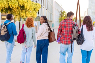 A grubu, genç ve çeşitli öğrenciler sırt çantalarıyla modern bir şehir bölgesinde yürüyorlar. Odak noktası, geriye bakan gülümseyen bir kadın. Sonbahar renkleriyle parlak bir atmosfer.