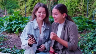 Siz iki neşeli genç bayan, dışarıda oturmuş, akıllı telefona bakarken bir anı paylaşıyorsunuz. Bereketli yeşilliklerle çevrili olan sahne, arkadaşlığı ve bağlantıyı gelişigüzel bir ortamda aktarıyor.