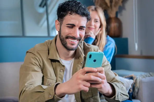 Güler yüzlü bir adam, neşeli bir adam evinde otururken elinde akıllı bir telefon tutuyor. Arka planda gülümseyen bir kadın onu ilgiyle izliyor..
