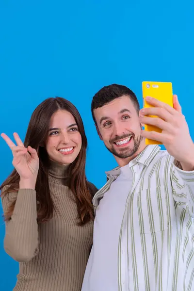  Neşeli genç bir çift, sarı akıllı telefon kullanarak selfie çekiyor. Kadın bir barış işareti gösterirken, adam parlak bir şekilde gülümsüyor. Parlak mavi arka plan canlı ve modern bir hava katıyor..
