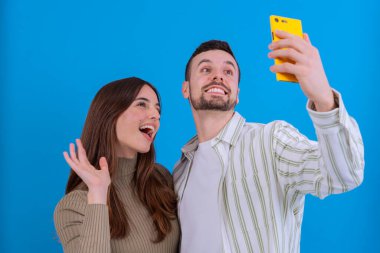  Neşeli genç bir çift, sarı akıllı telefon kullanarak selfie çekiyor. Kadın bir barış işareti gösterirken, adam parlak bir şekilde gülümsüyor. Parlak mavi arka plan canlı ve modern bir hava katıyor..
