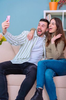  Neşeli genç bir çift, sarı akıllı telefon kullanarak selfie çekiyor. Kadın bir barış işareti gösterirken, adam parlak bir şekilde gülümsüyor. Parlak mavi arka plan canlı ve modern bir hava katıyor..