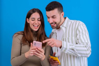  Neşeli bir çift rahat bir koltukta oturur, gülümser ve pembe bir akıllı telefonu işaret eder. Ev düzeninde bir kitap rafı ve çiçekler var, etkileşimleri için sıcak ve rahat bir atmosfer yaratıyorlar..