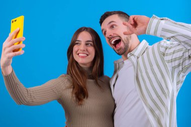 Genç çift, parlak mavi bir arka planda sarı bir akıllı telefonla selfie çekerken gülümsüyor. Sosyal medya, yaşam tarzı veya teknoloji temaları için ideal bir eğlence ve bağlantı anı yakalar. 