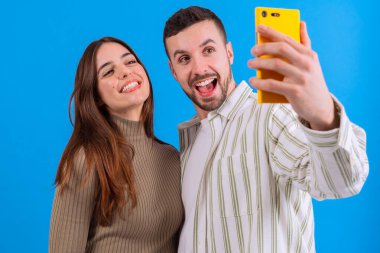 Genç çift, parlak mavi bir arka planda sarı bir akıllı telefonla selfie çekerken gülümsüyor. Sosyal medya, yaşam tarzı veya teknoloji temaları için ideal bir eğlence ve bağlantı anı yakalar. 