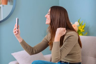 Neşeli bir kadın kanepede oturur, elinde akıllı bir telefonla sevinçle kutlama yapar. Arka planda çiçek dekoru olan rahat ev, sahneye canlı ve mutlu bir atmosfer katıyor..