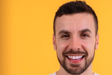 Cüretkar sarı bir arka planda parlak bir gülümsemesi olan neşeli bir adamın yakın çekimi. Canlı kompozisyon ve pozitif ifade onu yaşam tarzı, mutluluk ve reklam temaları için ideal kılıyor..