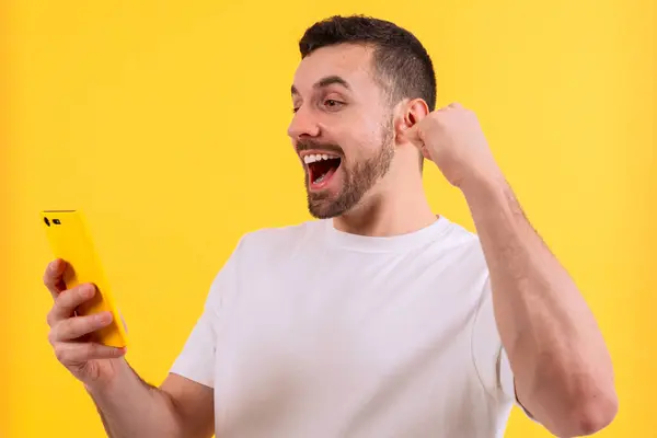 Heyecanlı neşeli bir adam sarı bir akıllı telefon tutuyor ve canlı sarı bir arka plana karşı heyecanla kutluyor. Başarı, teknoloji, mutluluk ve dijital yaşam tarzı içeriği temaları için mükemmel.