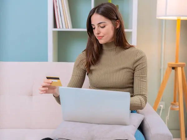 Neşeli bir kadın kanepede oturmuş internetten alışveriş yapıyor. Adamın elinde bir kredi kartı var, ikisi de heyecanlı bir şekilde dizüstü bilgisayara bakıyor.. 