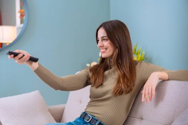 Bir kadın, elinde uzaktan kumandayla koltuğa rahatça oturur ve gülümser. Rahatlamış ve vaktinin tadını çıkarıyor. Parlak, rahat bir odası ve arka planda hafif ışıklandırması var..