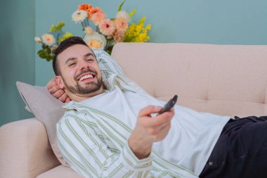 Rahatlamış bir adam gülümsüyor neşeli bir adam kanepede uzanıyor, elinde uzaktan kumandayla gülümsüyor. Arka planda dekoratif çiçekler olan rahat bir oturma odasında, sıcak bir atmosfer yaratıyor..